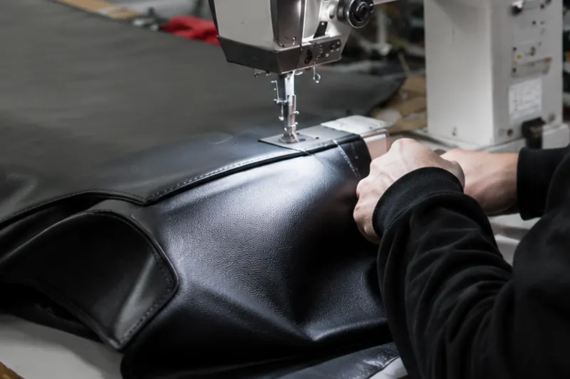 Schwarzes Lederteil unter dem Nähfuß einer Industrienähmaschine, zwei Hände führen den Zuschnitt
